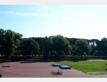 Campo sportivo all’aperto con pista di atletica in tartan che circonda un’area in erba. Sul lato si vede una pedana con materassi per salti e, sullo sfondo, una fila di alberi sotto un cielo sereno.