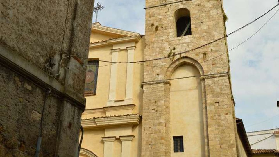 Campanile romanico in pietra con finestre ad arco sovrapposte e ingresso ad arco alla base, affiancato da una chiesa intonacata color chiaro. La torre si affaccia su una stretta piazza tra edifici in muratura, con cavi sospesi e cielo parzialmente nuvoloso sullo sfondo.