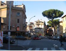 Incrocio urbano con strisce pedonali e auto parcheggiate lungo la strada, circondato da edifici residenziali di più piani con facciate in intonaco. Sullo sfondo si vede una scalinata che sale tra le palazzine e un grande pino mediterraneo vicino al margine destro.