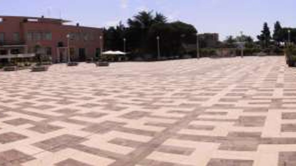 Ampia piazza pedonale pavimentata con motivi geometrici a riquadri chiari e scuri, con dissuasori in cemento e lampioni. Sullo sfondo si vedono edifici moderni in mattoni rossi, alcune aiuole e una struttura coperta con teli bianchi.
