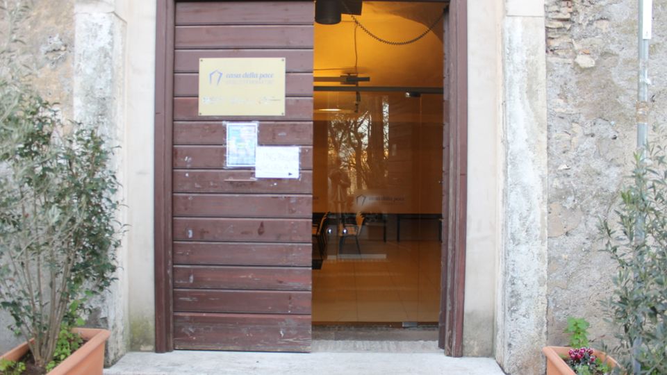 Ingresso di un edificio in pietra con grande porta in legno scorrevole e apertura che mostra un interno illuminato con pavimento piastrellato e pareti chiare. Ai lati dell’entrata ci sono vasi rettangolari con piante e una targa informativa affissa sulla porta.