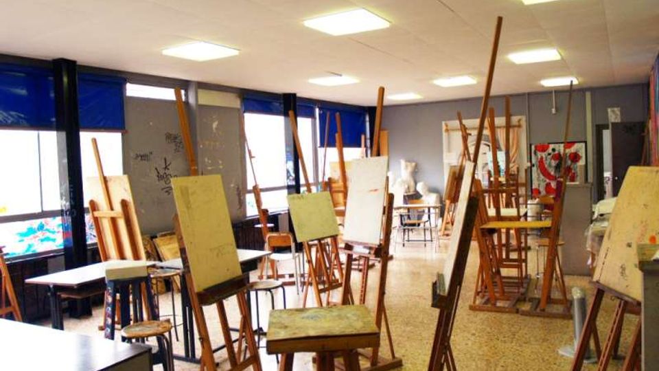 Aula di arte con numerosi cavalletti in legno e tele disposte tra tavoli e sgabelli su pavimento in graniglia. Grandi finestre laterali con tendine blu e illuminazione a pannelli sul soffitto.