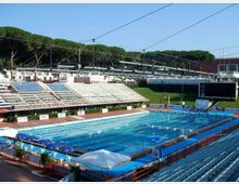 Piscina olimpionica all’aperto con corsie delimitate e trampolini, circondata da tribune con gradinate e sedute. Sullo sfondo si vedono una grande schermata elettronica, strutture metalliche con cavi e un’area verde con alberi.