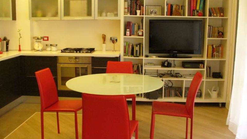 Interno open space con cucina moderna ad angolo, pensili con ante in vetro e piano di lavoro chiaro con forno e fornelli integrati. In primo piano c’è un tavolo rotondo con quattro sedie rosse, e sullo sfondo una grande libreria bianca con TV e scaffali pieni di libri e oggetti.