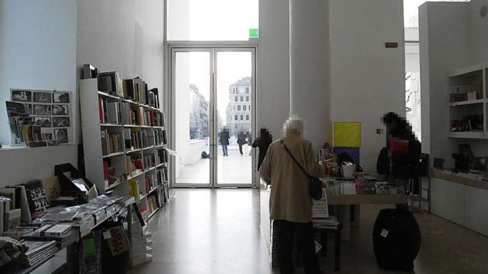 Interno di un negozio di libri o spazio espositivo moderno con soffitti alti, scaffali e tavoli pieni di volumi e riviste. In fondo, una grande porta vetrata a tutta altezza si apre su una piazza esterna con edifici visibili.