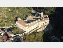 Veduta aerea di un grande complesso monastico in pietra con tetti in coppi, chiostro interno e torre campanaria, affacciato su un pendio. Intorno si vedono strade, case del borgo e campi coltivati con alberi in un’area collinare boscosa.