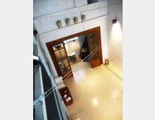 Interno di una galleria o museo visto dall’alto, con pavimento chiaro a piastrelle e grandi porte in legno con pannelli vetrati e decorati che conducono a una sala illuminata. Sulla parete superiore sono allineati busti decorativi e l’ambiente è illuminato da faretti, con un tavolo e piccoli arredi lungo il perimetro.