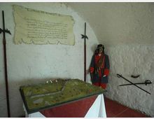 Piccola sala espositiva con pareti bianche in muratura, con una vetrina/maquette di terreno su un tavolo al centro e un cartiglio storico appeso alla parete. Alle pareti sono esposti attrezzi e armi antiche, tra cui una lunga alabarda e una bilancia, con una figura in uniforme d’epoca nell’angolo.