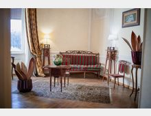 Salotto in stile classico con pavimento in parquet e tappeto, divano e sedie imbottite a righe rosse e verdi, tavolino in legno e lampade su alti piedistalli intagliati. Una grande finestra con tende pesanti illumina la stanza, con vasi decorativi e un quadro appeso alla parete.