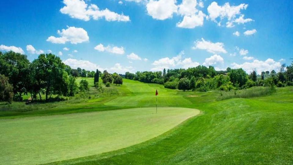 Campo da golf con ampio green in primo piano e bandierina centrale, circondato da colline erbose e alberi. Sullo sfondo cielo azzurro con nuvole sparse.