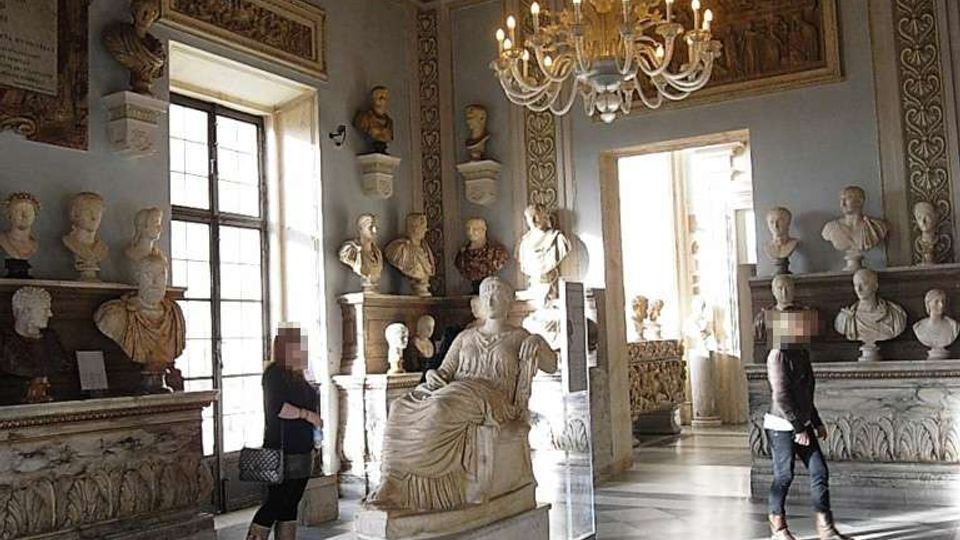 Interno di una sala museale con pavimento in marmo a motivi geometrici, pareti ornate da rilievi e busti scultorei, e una statua seduta su piedistallo al centro. Dal soffitto a cassettoni decorato pendono grandi lampadari, e ampie finestre e porte fanno entrare luce naturale.
