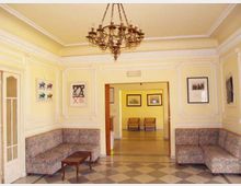 Salotto o area d’attesa in stile classico con pareti gialle decorate da cornici in gesso, lampadario centrale e divani angolari con tavolino. Un’ampia apertura conduce a una stanza adiacente con sedie e quadri, su pavimento a scacchi.