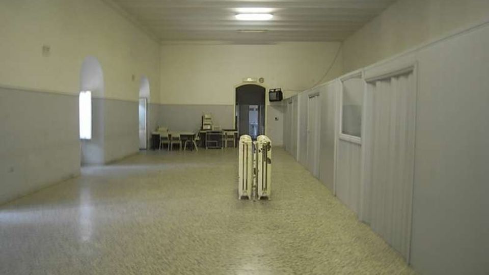 Lungo corridoio interno con pavimento in graniglia e pareti bicolori, illuminato da plafoniere a soffitto. Sul lato destro si vedono pareti divisorie con porte e finestre, mentre in fondo ci sono alcune sedie e tavoli vicino a una porta ad arco.