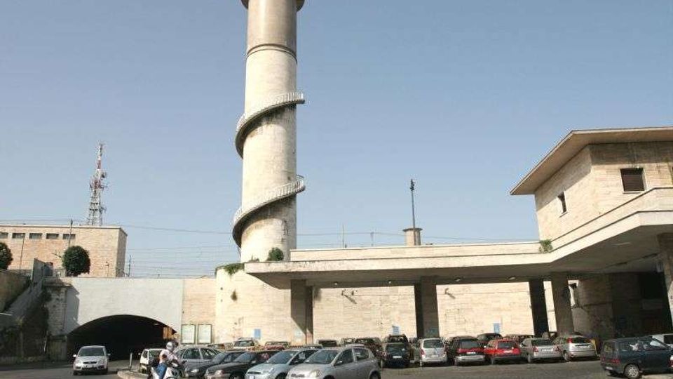 Parcheggio esterno di un complesso industriale in pietra chiara, con una alta torre cilindrica dotata di scala a spirale esterna. In primo piano sono visibili numerose auto parcheggiate, con un portico lungo e un’ampia apertura ad arco che conduce a un’area coperta.