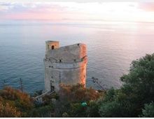 Torre costiera in pietra su un promontorio roccioso, circondata da vegetazione mediterranea e affacciata sul mare calmo. Sullo sfondo si vede l’orizzonte marino con cielo al tramonto.