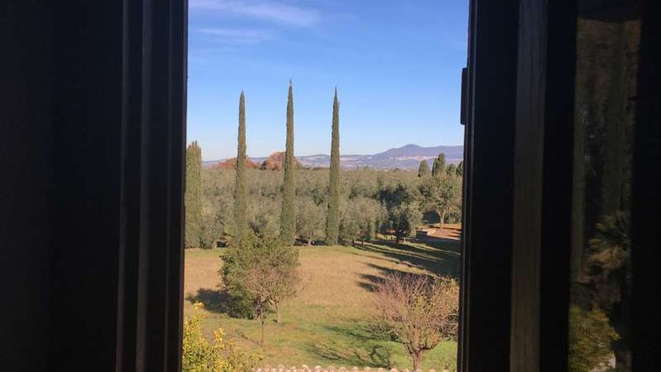 Vista attraverso una finestra rettangolare in un interno scuro, che incornicia un paesaggio di campagna con prato, alberi e alti cipressi. Sullo sfondo si vedono colline o montagne sotto un cielo sereno.