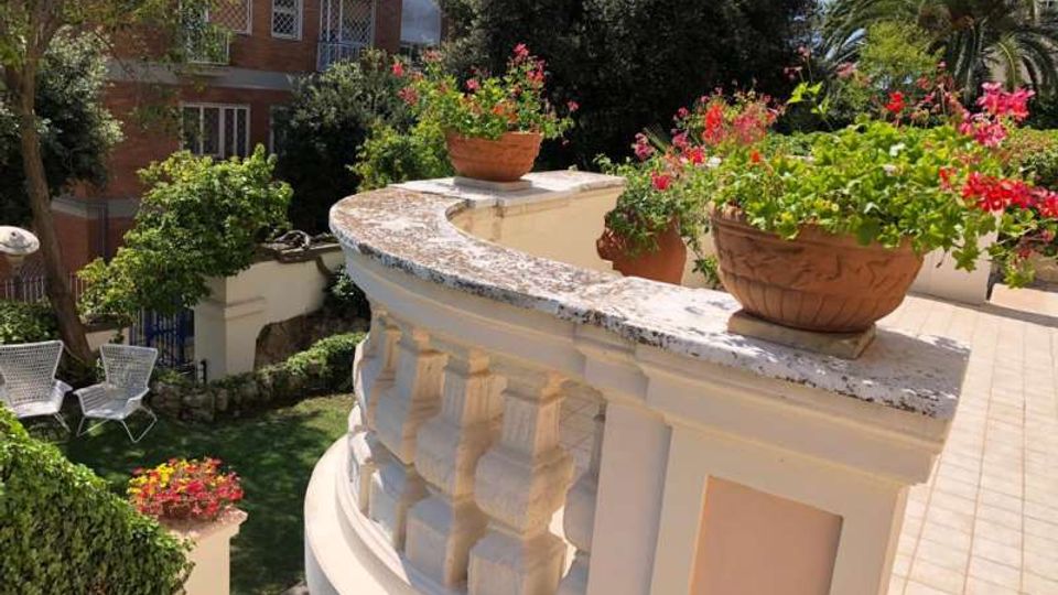 Terrazza esterna con balaustra curva in pietra chiara e vasi di terracotta con fiori rossi, affacciata su un giardino verde sottostante. Sullo sfondo si vedono alberi e palme e un edificio residenziale con balconi.