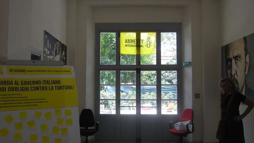 Atrio interno con soffitto a pannelli e una grande porta a vetri che affaccia su alberi all’esterno, con uno striscione giallo “Amnesty International” sopra l’ingresso. Ai lati si vedono un pannello informativo e un tavolo con materiali e volantini su tovaglia gialla.