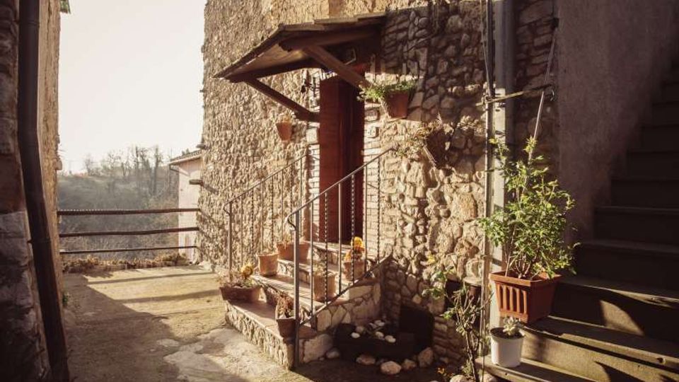 Cortile esterno in un borgo, con edificio in pietra e piccola veranda sopra l’ingresso raggiunto da pochi gradini con ringhiera in ferro. Vasi di piante lungo il muro e su una scala laterale, con parapetto sullo sfondo affacciato su una valle.
