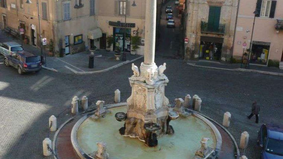 Fontana monumentale al centro di una piazza lastricata, con grande vasca circolare e colonna in pietra decorata con sculture. Intorno si vedono palazzi storici dai toni pastello con tetti in tegole e una strada stretta che si apre tra gli edifici.