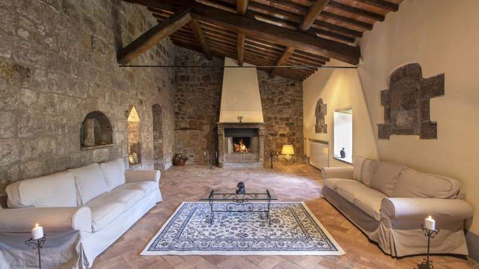 Interno di un soggiorno rustico con pareti in pietra a vista, soffitto con travi in legno e pavimento in cotto. Al centro c’è un camino acceso con ampia cappa, affiancato da due divani chiari, un tappeto decorato e un tavolino in ferro, con nicchie e finestre incassate nelle mura.