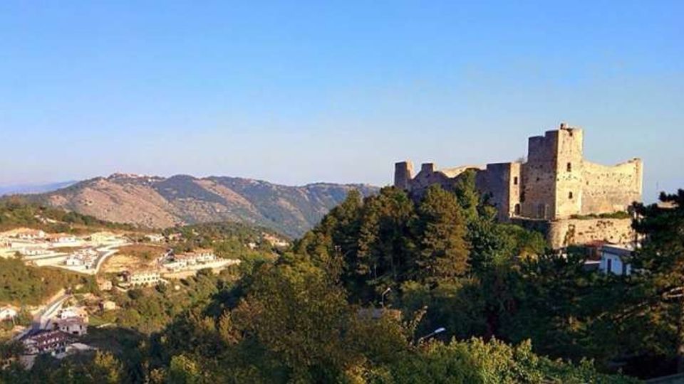 Panorama collinare con una grande fortezza in pietra in cima a un’altura, circondata da boschi e alberi. Sullo sfondo si vedono montagne e un piccolo centro abitato con case sparse lungo la valle sotto un cielo sereno.