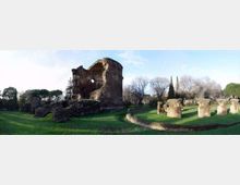 Panorama di un’area archeologica con grandi rovine in mattoni di un edificio antico e diversi resti di muri e pilastri sparsi su un prato. Sullo sfondo si vedono alberi e cipressi sotto un cielo sereno.