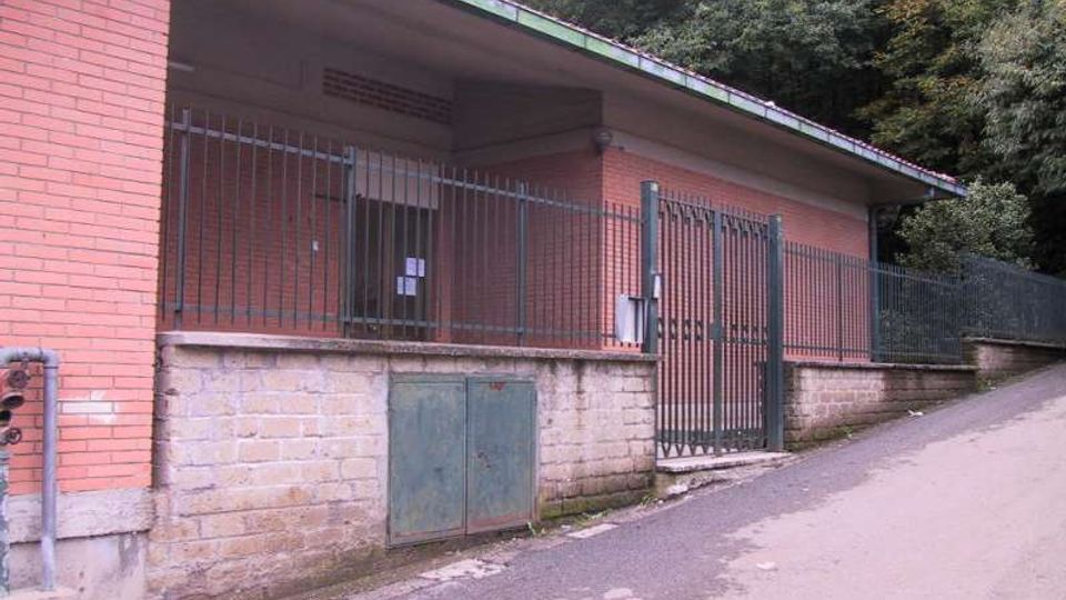 Edificio basso in mattoni con tetto a falda e recinzione metallica, affiancato a una strada in salita. Alla base c’è un muretto in cemento con sportello metallico, sullo sfondo una fitta vegetazione boschiva.