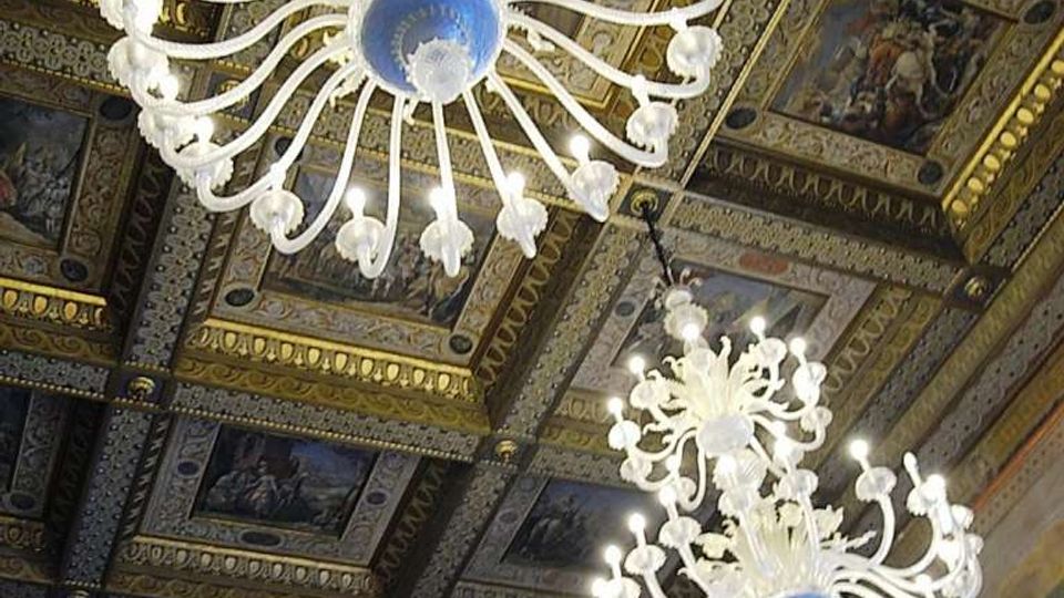 Soffitto di una sala storica riccamente decorato con riquadri dipinti e cornici dorate, con grandi lampadari in vetro bianco sospesi. Sulle pareti sottostanti si intravedono affreschi e archi in stile classico.