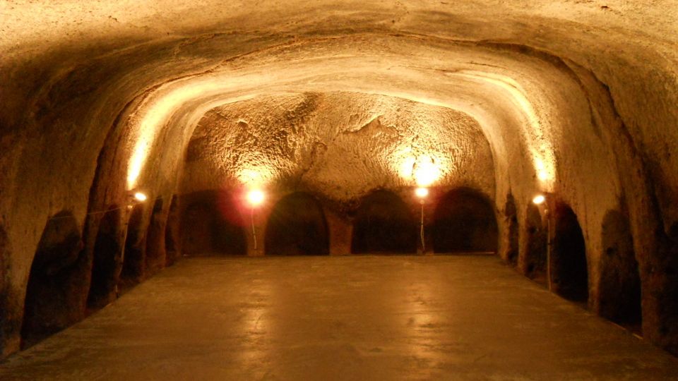 Interno di una grotta o cantina sotterranea con soffitto a volta scavato nella roccia e pavimento in cemento. Luci calde lungo le pareti illuminano una serie di nicchie e archi sul fondo.