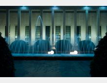 Fontana illuminata di notte con getti d’acqua davanti a un edificio monumentale con alte colonne e grandi finestre verticali. Nella vasca si vedono sfere luminose bianche e siepi potate, con un’ampia pavimentazione in primo piano.
