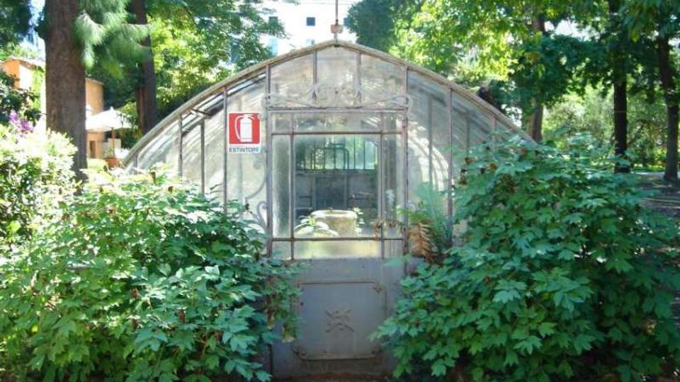 Piccola serra con struttura ad arco in metallo e vetro, con porta centrale e cartello “Estintore”, situata in un parco alberato. Fitta vegetazione e cespugli circondano l’ingresso, con un vialetto in primo piano.
