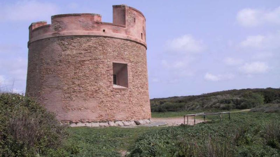 Torre cilindrica in mattoni con sommità merlata e un’apertura rettangolare, situata in una zona di campagna con vegetazione bassa e sentiero sterrato. Sullo sfondo si vedono colline verdi sotto un cielo parzialmente nuvoloso.