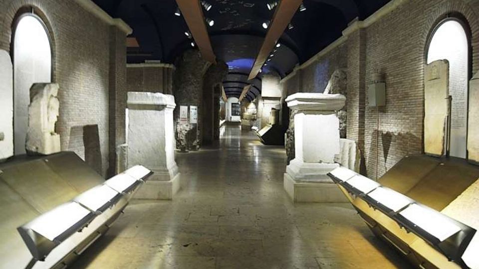 Interno di un corridoio museale lungo e simmetrico con pareti in mattoni e soffitto a volta scuro, illuminato da faretti. Ai lati si vedono basamenti e reperti in pietra, pannelli espositivi inclinati e grandi aperture ad arco incassate nelle pareti.
