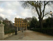 Viale pedonale in un parco urbano con percorso in ghiaia, siepi e grandi alberi spogli ai lati. A sinistra ci sono una recinzione in ferro con muretto e lampioni, sullo sfondo un edificio giallo a più piani sotto cielo nuvoloso.