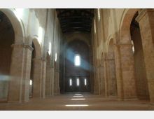 Interno di una chiesa in stile romanico con navata centrale ampia, file di pilastri in pietra e arcate laterali. Piccole finestre ad arco sul fondo lasciano entrare fasci di luce che illuminano il pavimento.