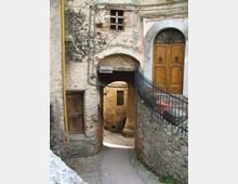 Stretto vicolo in pietra di un borgo antico, con passaggio ad arco sotto un edificio intonacato e scrostato e una piccola finestra con grata. Sulla destra si vede un portone in legno con arco superiore e una ringhiera in ferro lungo un muretto di pietra.