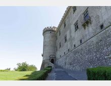 Veduta esterna di un castello in pietra con mura alte e una torre cilindrica merlata, con piccole finestre e un balconcino. Un vialetto in salita costeggiato da siepi conduce a un portale d’ingresso, con prato e alberi sul lato sinistro.