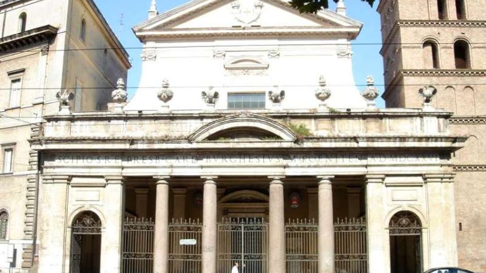 Facciata di una chiesa monumentale in stile classico con portico a colonne e cancellata in ferro, sormontata da un frontone triangolare. Ai lati si vedono edifici adiacenti, tra cui un campanile in mattoni, lungo una strada urbana.