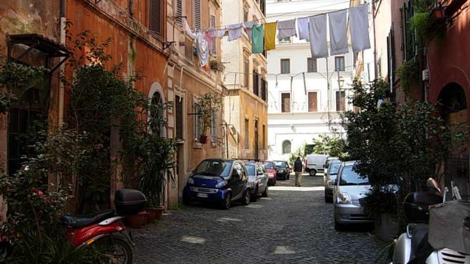 Vicolo urbano stretto con pavimentazione in sampietrini, fiancheggiato da edifici alti con facciate color ocra e persiane. Auto e scooter parcheggiati lungo i lati, piante in vaso e panni stesi su fili tesi tra i palazzi in fondo alla strada.