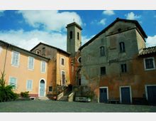 Piccola piazza lastricata con edifici in intonaco ocra e grigio e una chiesa affiancata da un campanile quadrato con croce in cima. Si vedono scale in pietra che salgono all’ingresso e facciate con finestre e porte sotto un cielo azzurro con nuvole.
