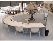 Sala di biblioteca o studio moderna con grande tavolo circolare bianco e sedie, al centro un globo terrestre antico su supporto decorato. Sullo sfondo ci sono lunghe finestre con tende, tavoli in legno e scaffali pieni di libri.