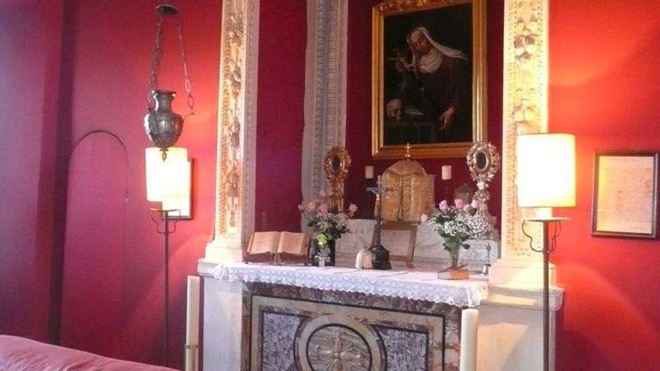 Interno di una piccola cappella con pareti rosso scuro e altare in marmo decorato, incorniciato da stucchi bianchi e sormontato da un dipinto religioso. Sul banco dell’altare sono presenti candelabri, fiori e libri, con lampade a parete e una lampada sospesa che illuminano la scena.