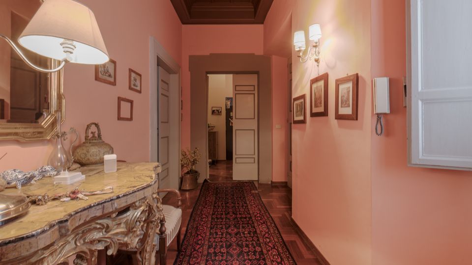Corridoio interno di una casa con pareti rosa e soffitto a cassettoni scuro, pavimento in parquet e lungo tappeto persiano centrale. A sinistra c’è una consolle decorata con grande specchio e lampada, mentre a destra si vedono applique, quadretti incorniciati e un citofono vicino a una finestra; in fondo una porta conduce ad altre stanze.