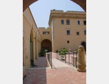 Cortile interno con pavimentazione in mattoni e passaggio centrale delimitato da una ringhiera metallica, affiancato da un edificio storico giallo con arcate e finestre. In primo piano si vede una colonna o statua in pietra e sullo sfondo un portico con grande arco che conduce a un’area ombreggiata.