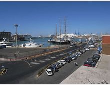 Veduta di un porto con banchine e parcheggi, una strada a più corsie in primo piano e diverse imbarcazioni ormeggiate, tra cui un grande veliero con alberi alti. Sullo sfondo si vedono navi più grandi e gru portuali sotto un cielo sereno.