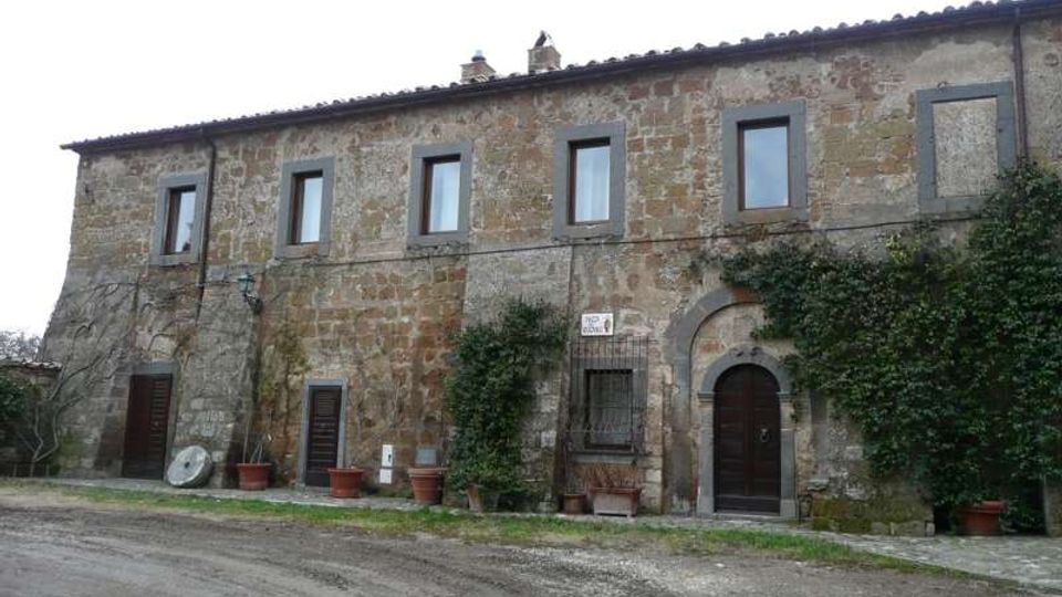 Facciata di un casale rurale in pietra a due piani, con tetto in coppi e finestre rettangolari incorniciate. Al piano terra ci sono porte in legno, un portale ad arco e piante rampicanti con grandi vasi lungo la base, affacciati su una strada sterrata.