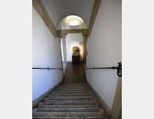 Scala interna in pietra che scende verso un corridoio con pavimento in mattoni e pareti bianche, dotata di corrimano in ferro ai lati. In fondo si vede un arco in pietra e una scultura di leone su piedistallo, illuminata da una luce a parete.