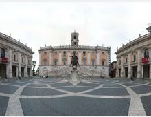 Ampia piazza lastricata con disegni geometrici, dominata da una statua equestre centrale e da un palazzo storico con facciata rosa, scalinata e torre dell’orologio. Ai lati si vedono due edifici porticati simmetrici che incorniciano lo spazio aperto.