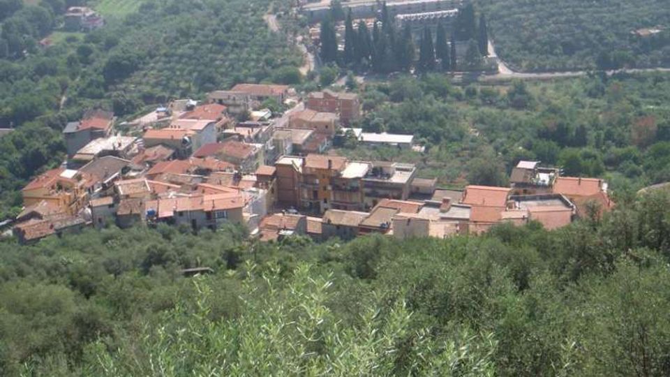 Veduta dall’alto di un piccolo borgo collinare con case addossate e tetti in coppi rossi, circondato da uliveti e vegetazione fitta. Sullo sfondo si vede una strada sinuosa e un complesso di edifici immerso nel verde con alti cipressi.